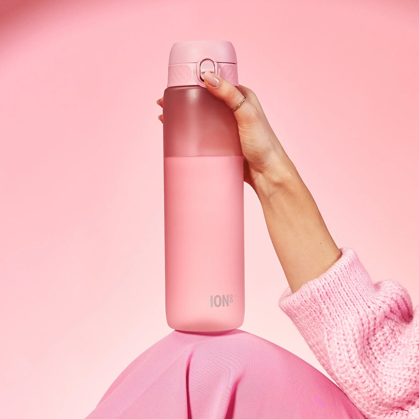 A hand holds a pink water bottle labeled ION8 against a pink background while wearing a pink sweater