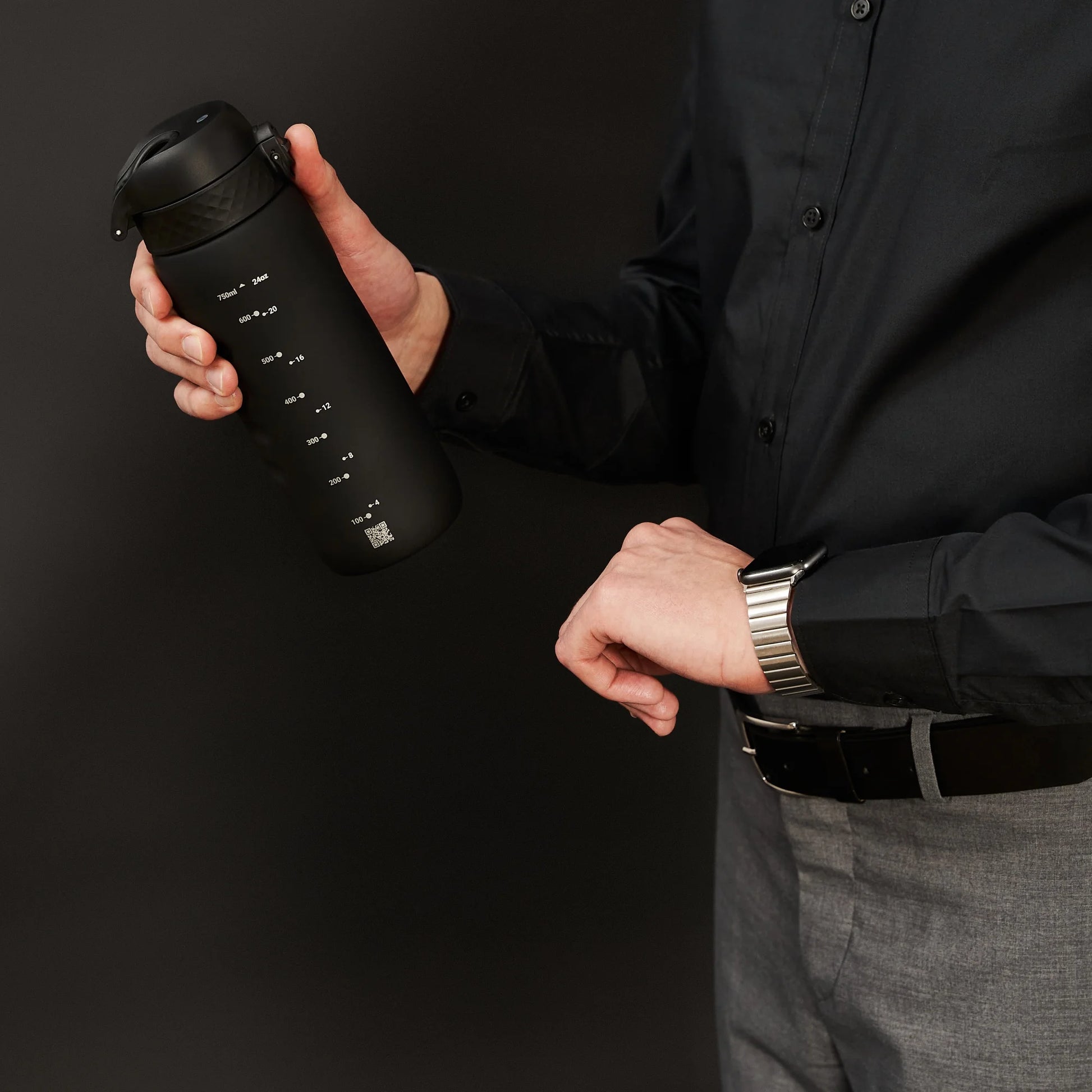 A person holds a black water bottle with measurement markings and a QR code while adjusting their smartwatch in a formal setting wearing a black shirt and gray pants.