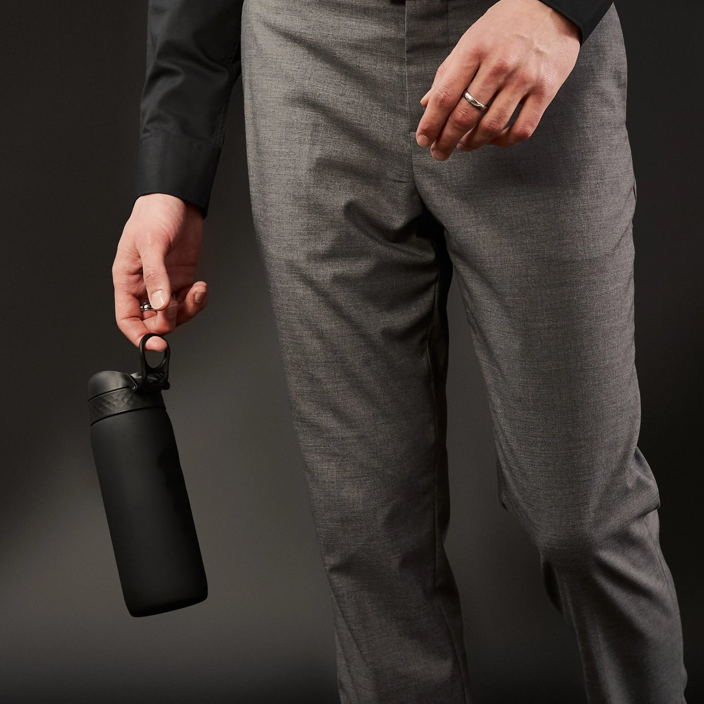 A person holds a black water bottle by its loop in their left hand wearing a dark shirt and gray pants against a dark background