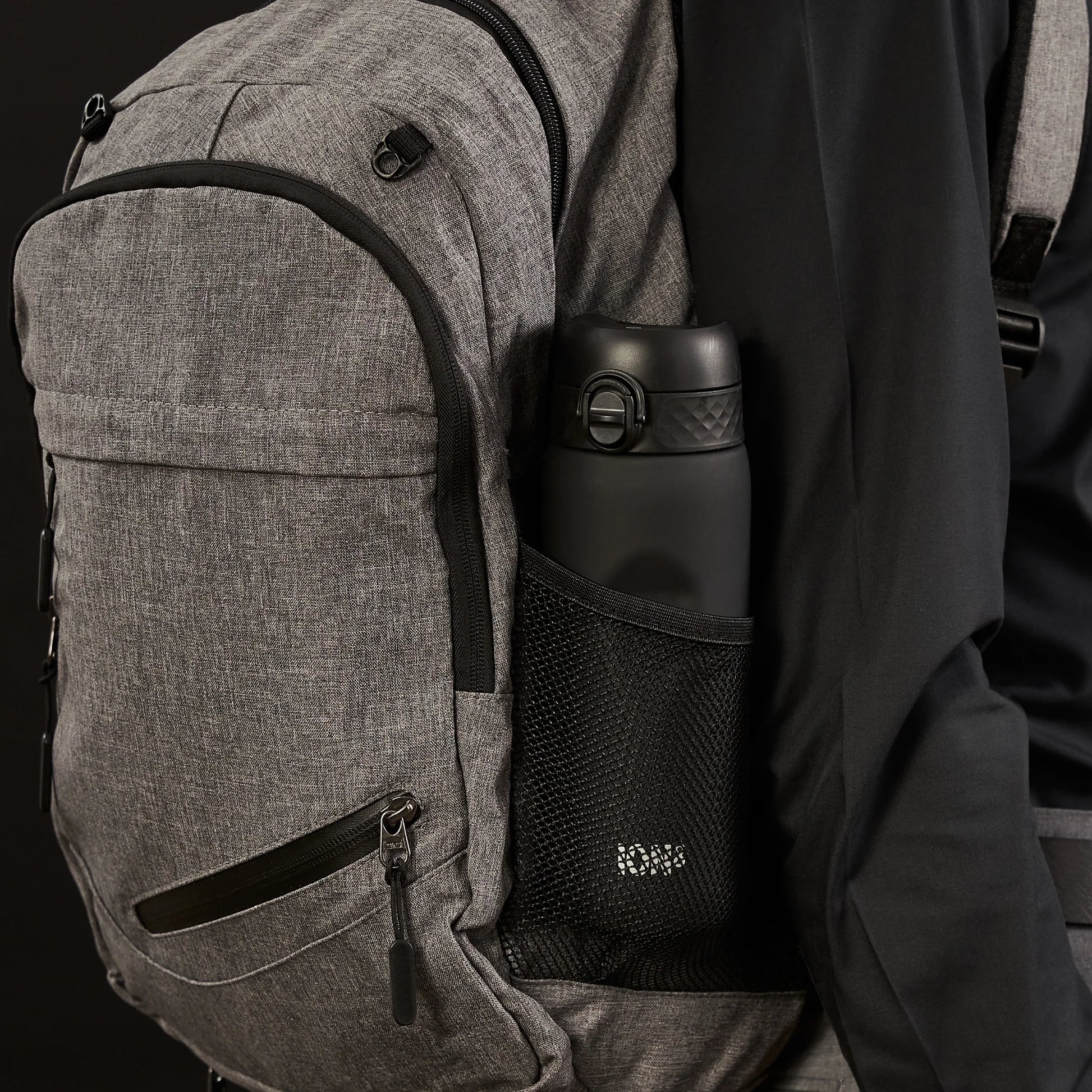 A gray backpack holds a black water bottle in its side mesh pocket against a dark background.