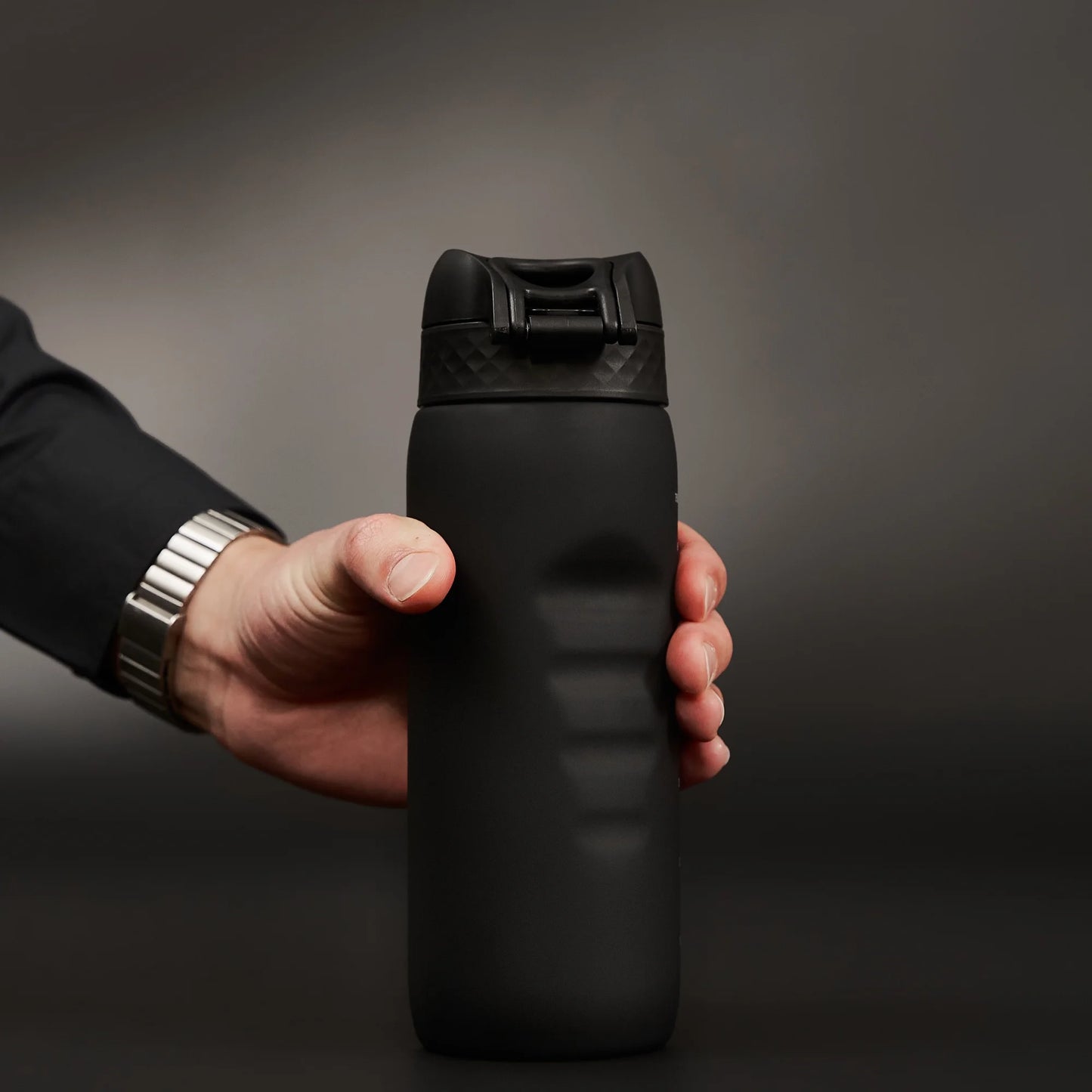 A person holds a black insulated water bottle with a textured cap against a neutral gray background. The bottle is gripped firmly, and the hand wears a silver watch.