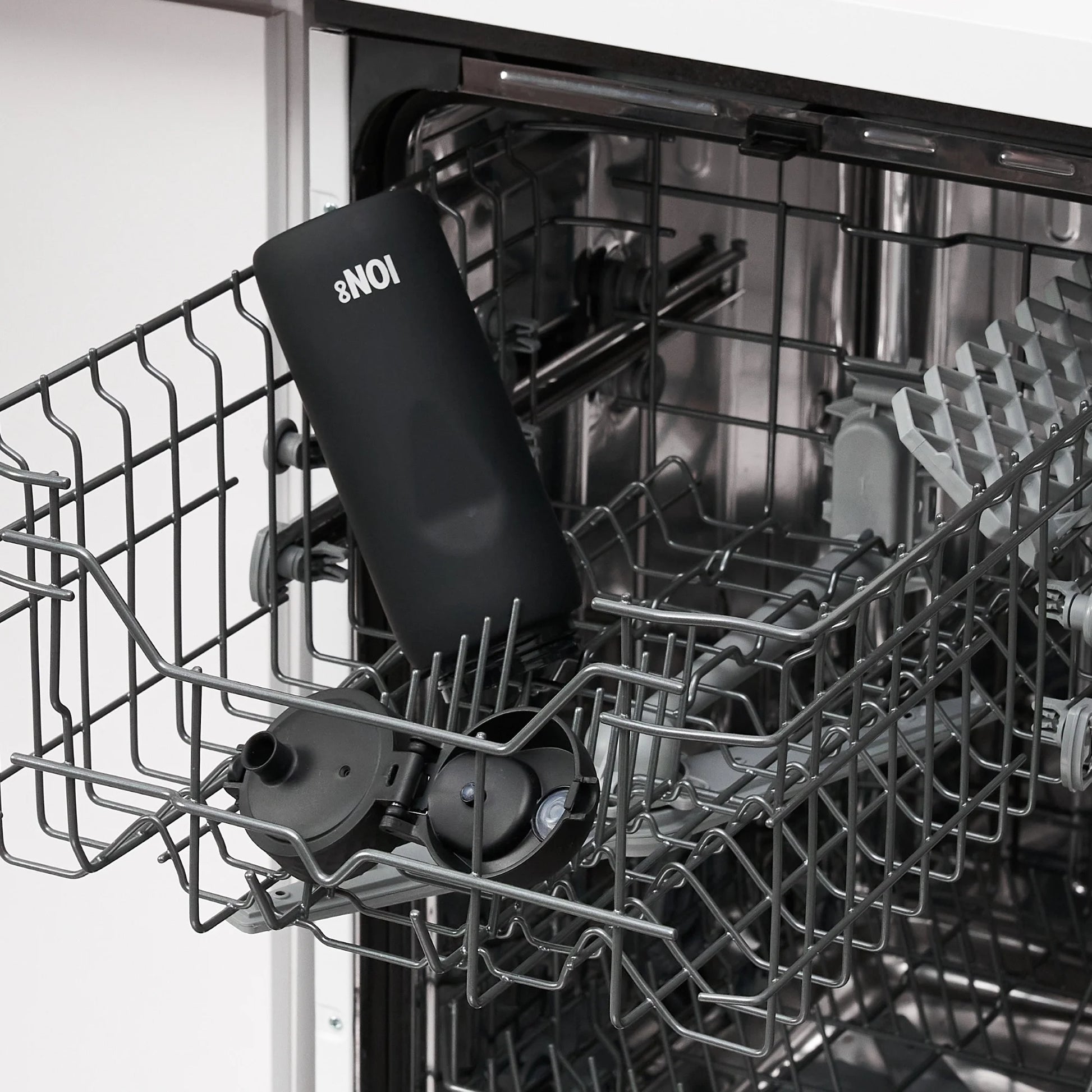 A black bottle labeled "NO18" rests upside down in an open dishwasher rack surrounded by metal prongs and kitchen utensils inside a modern kitchen setting.