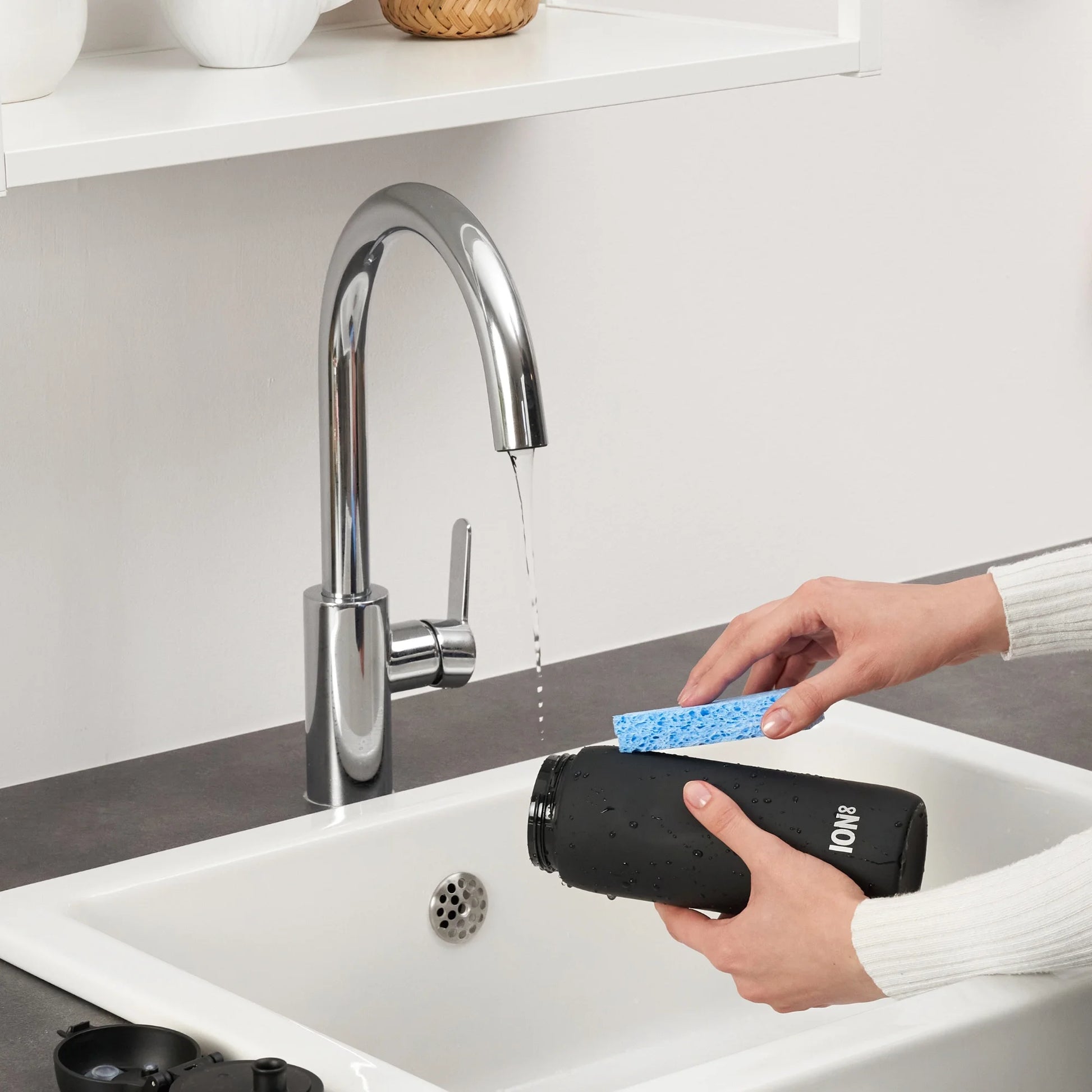 Hands clean a black water bottle with a blue sponge under a running kitchen faucet in a modern, minimalist kitchen setting. The bottle has "ION8" printed on it.