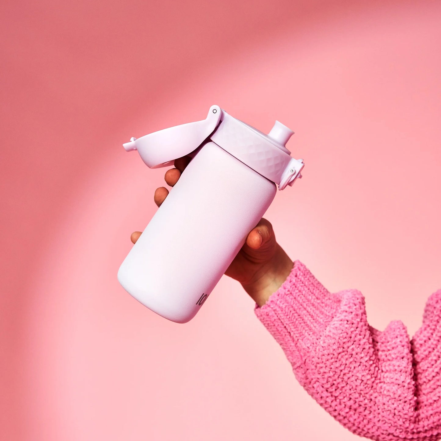 A hand in a pink sweater holds a pink insulated bottle with a flip lid against a pink background.