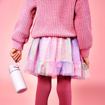 A child in a pink sweater and tutu holds a white water bottle against a pink background.