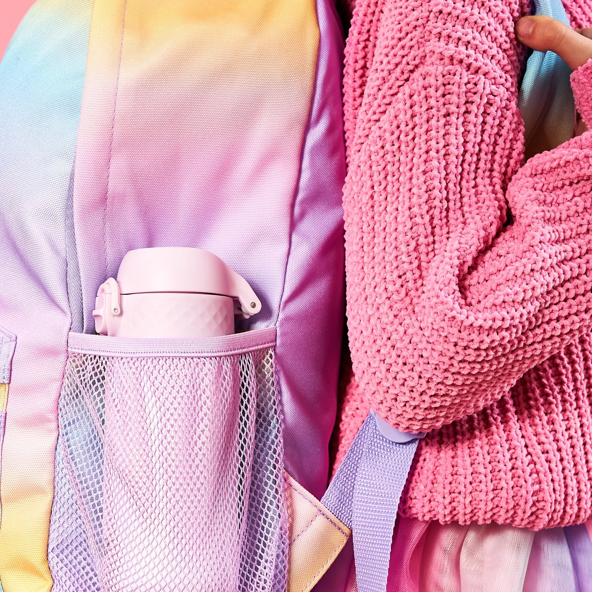 Backpack holds a pink water bottle in a mesh side pocket while a person in a pink sweater carries it against a pastel background