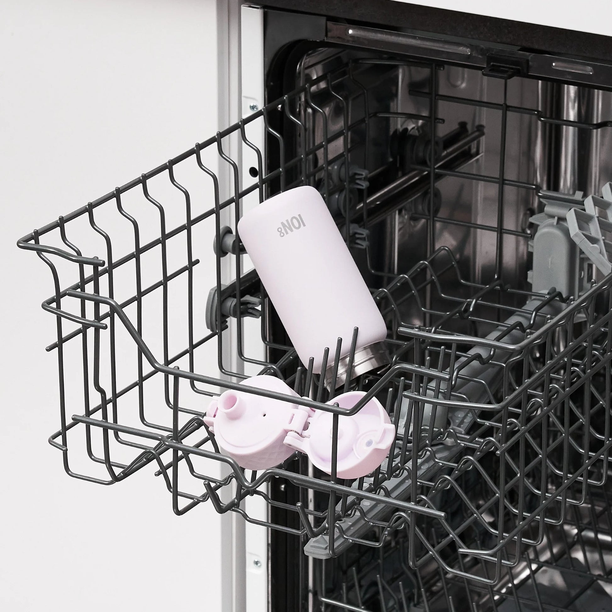 A white bottle labeled "8NOI" and a pink lid are placed inside an open dishwasher rack in a kitchen environment.