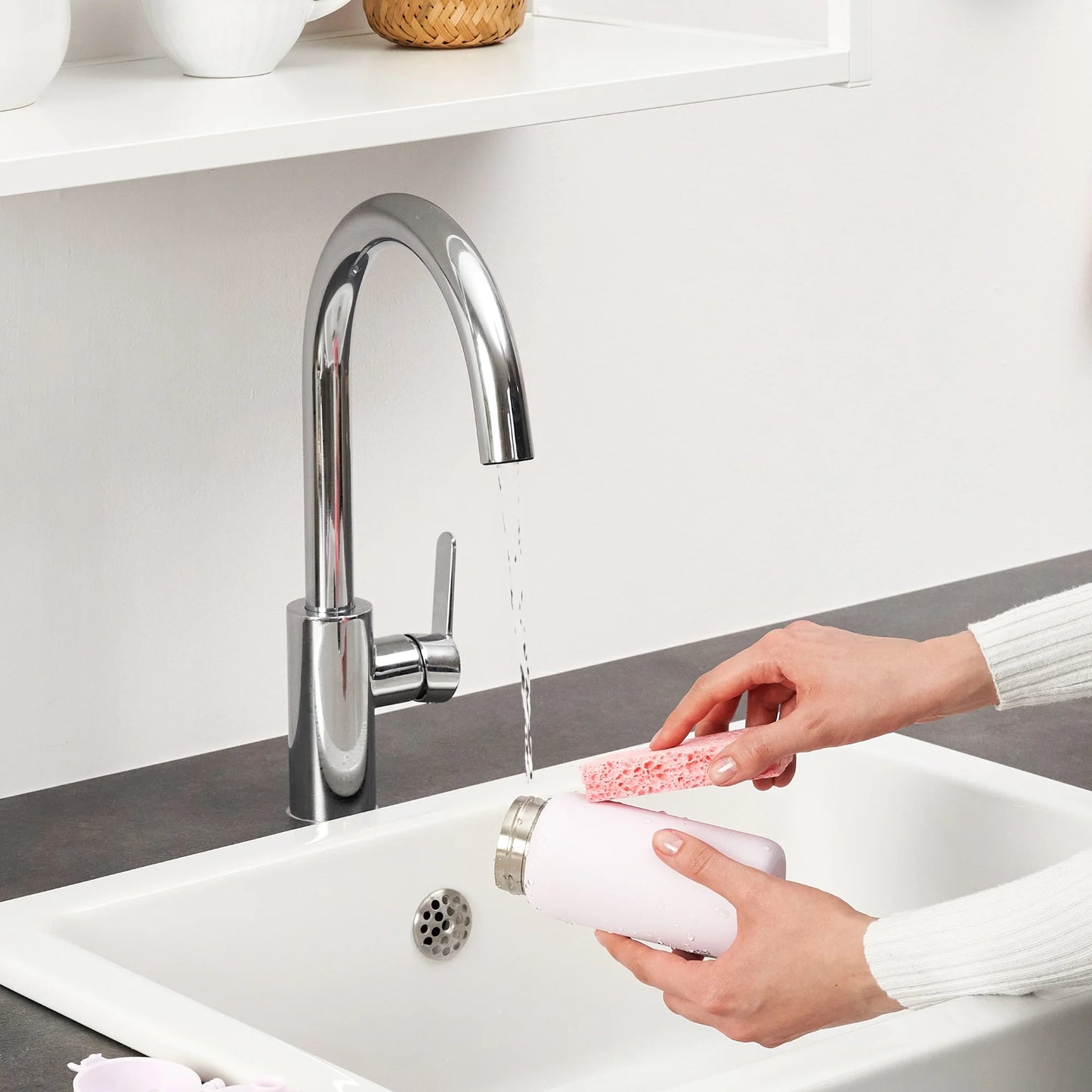 Hands scrubbing a white bottle with a pink sponge under running water in a modern kitchen sink surrounded by white shelves and décor items.