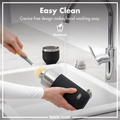 A hand scrubs a black travel flask with a brush under a running faucet in a kitchen sink. Text: Easy Clean Crevice free design makes hand washing easy. Handwash TRAVEL FLASK.