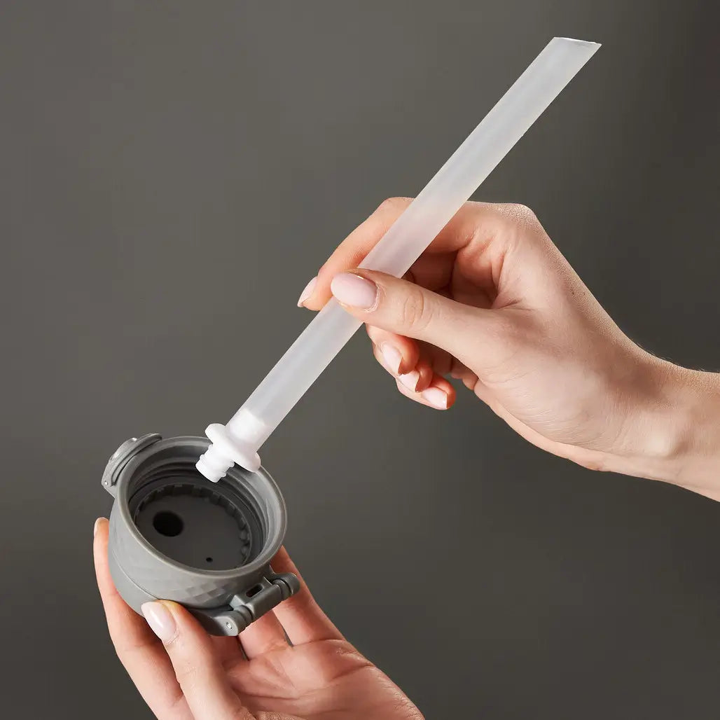 A gray bottle cap is being fitted with a translucent straw by a person's hands against a gray background.