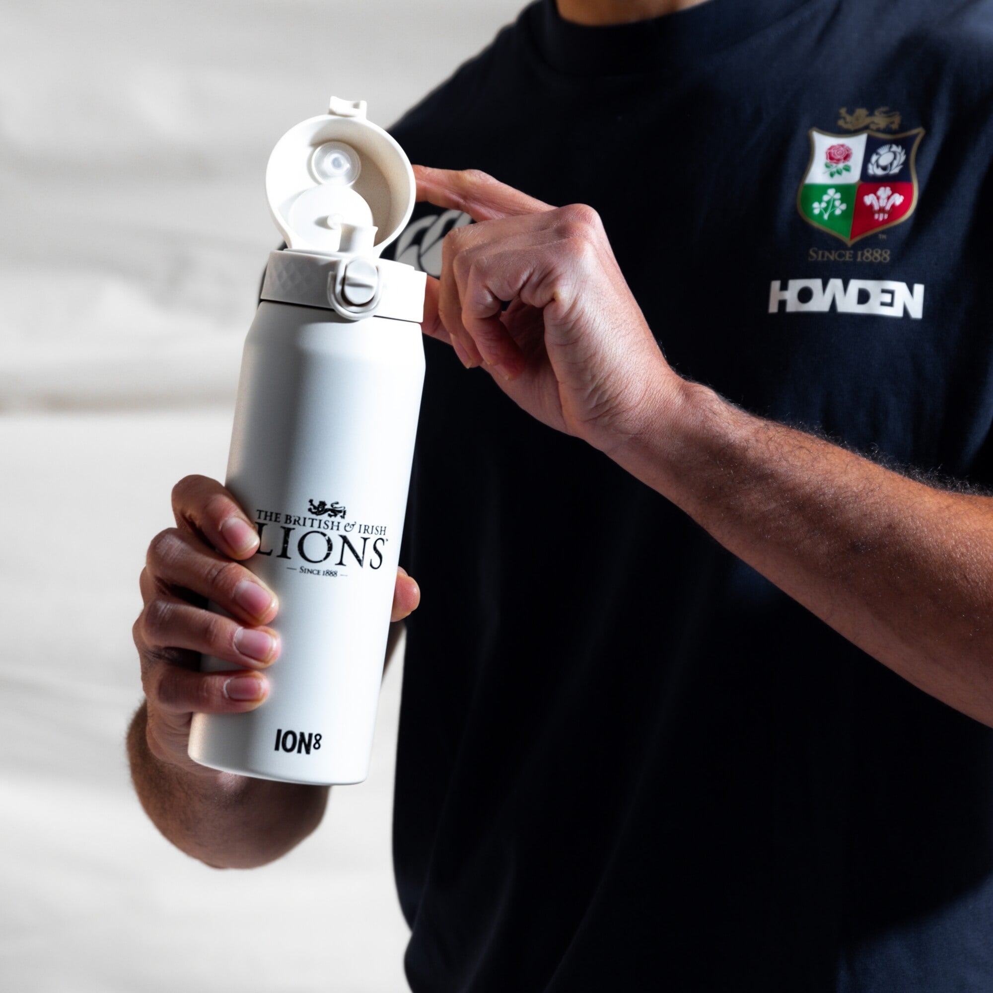 A white ION8 water bottle, featuring "THE BRITISH & IRISH LIONS - SINCE 1888," is being opened by a person wearing a black Howden rugby shirt.