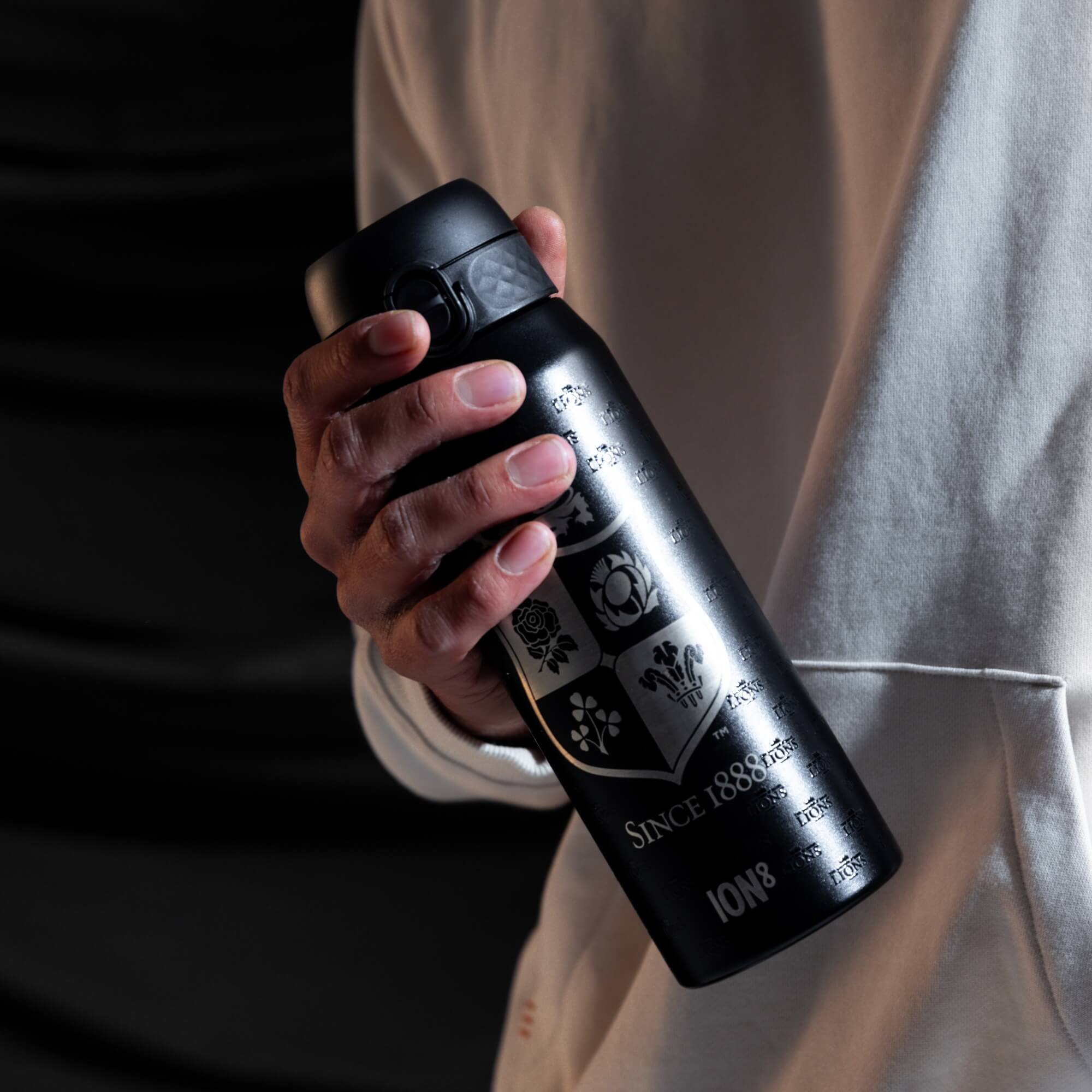 A black, cylindrical water bottle with engraved crests and "SINCE 1888" and "ION8" is held; the hand and bottle are against a dark background and light-colored clothing.