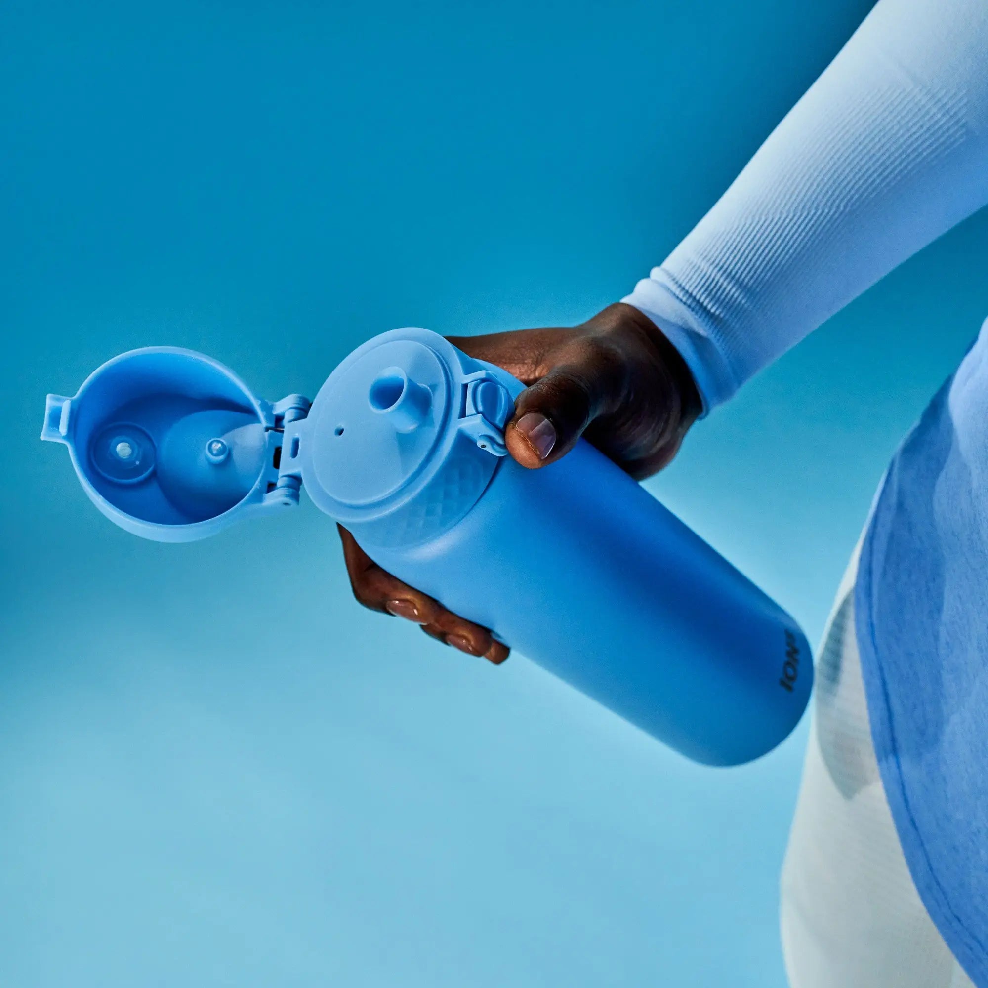A hand holds an open blue water bottle with a flip-top lid against a blue background wearing a long-sleeved shirt