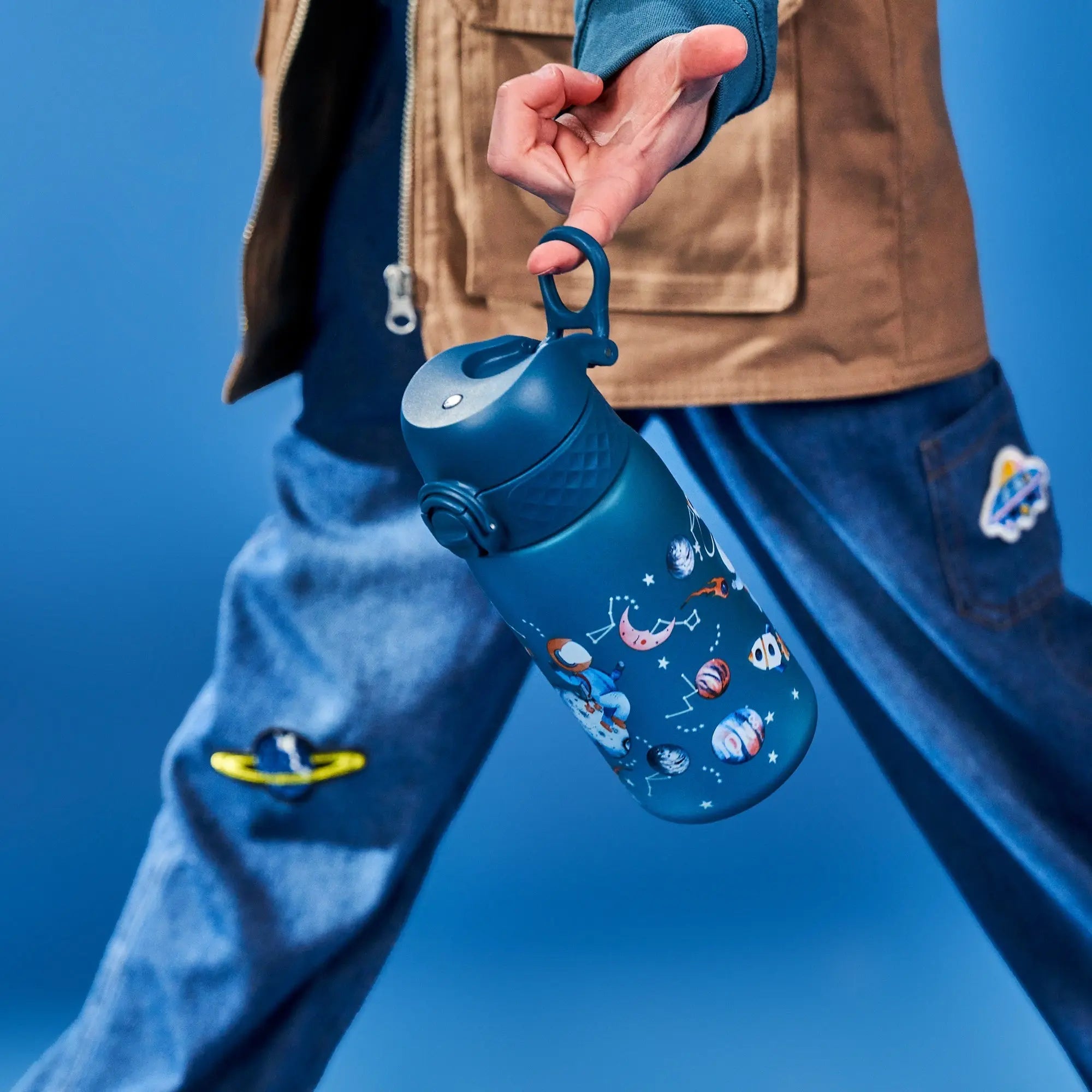 A person carries a blue water bottle with space-themed illustrations, wearing blue jeans and a tan jacket, set against a blue background.