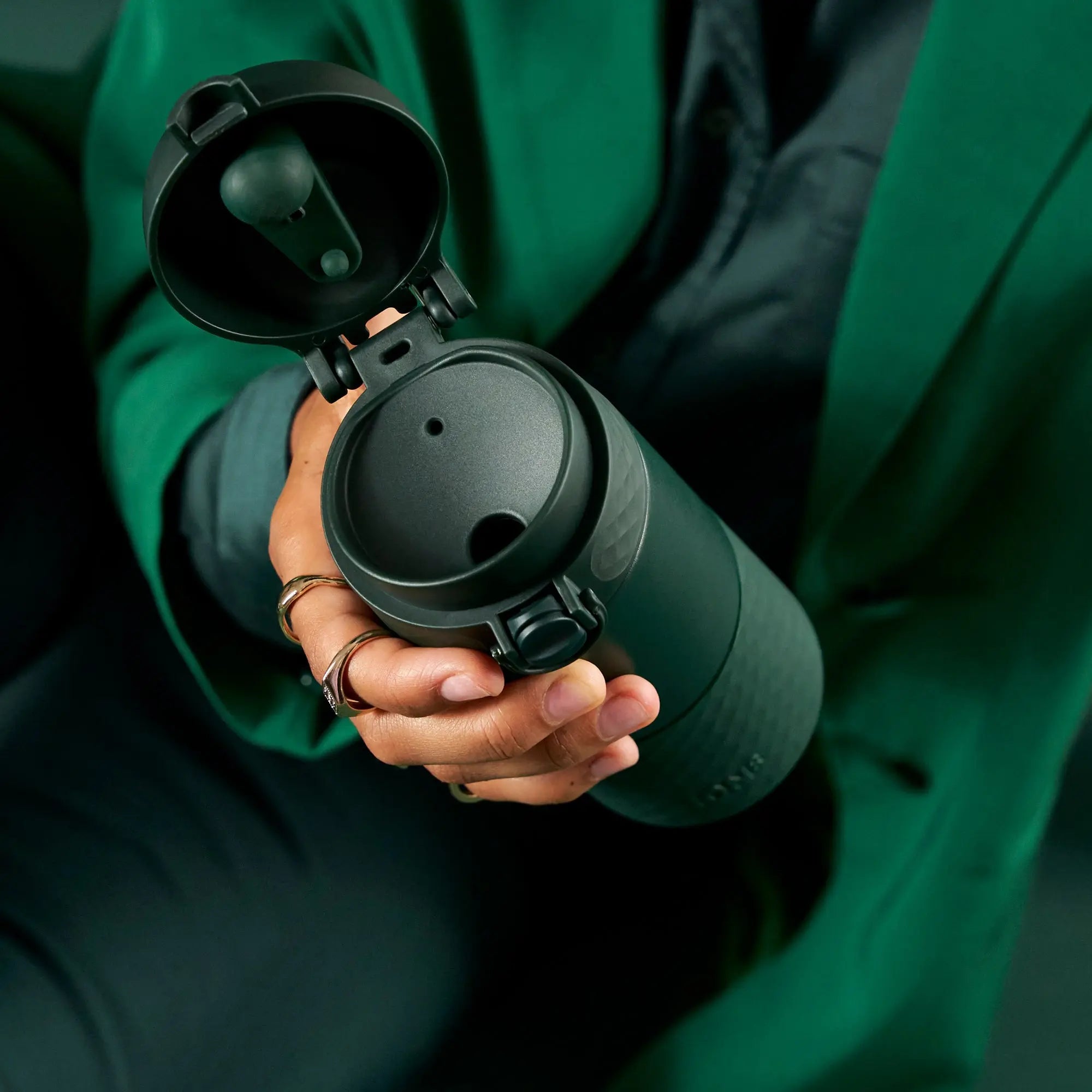 A person holds an open black insulated travel mug with a flip-top lid in their hand wearing rings and a green jacket in a casual indoor setting.