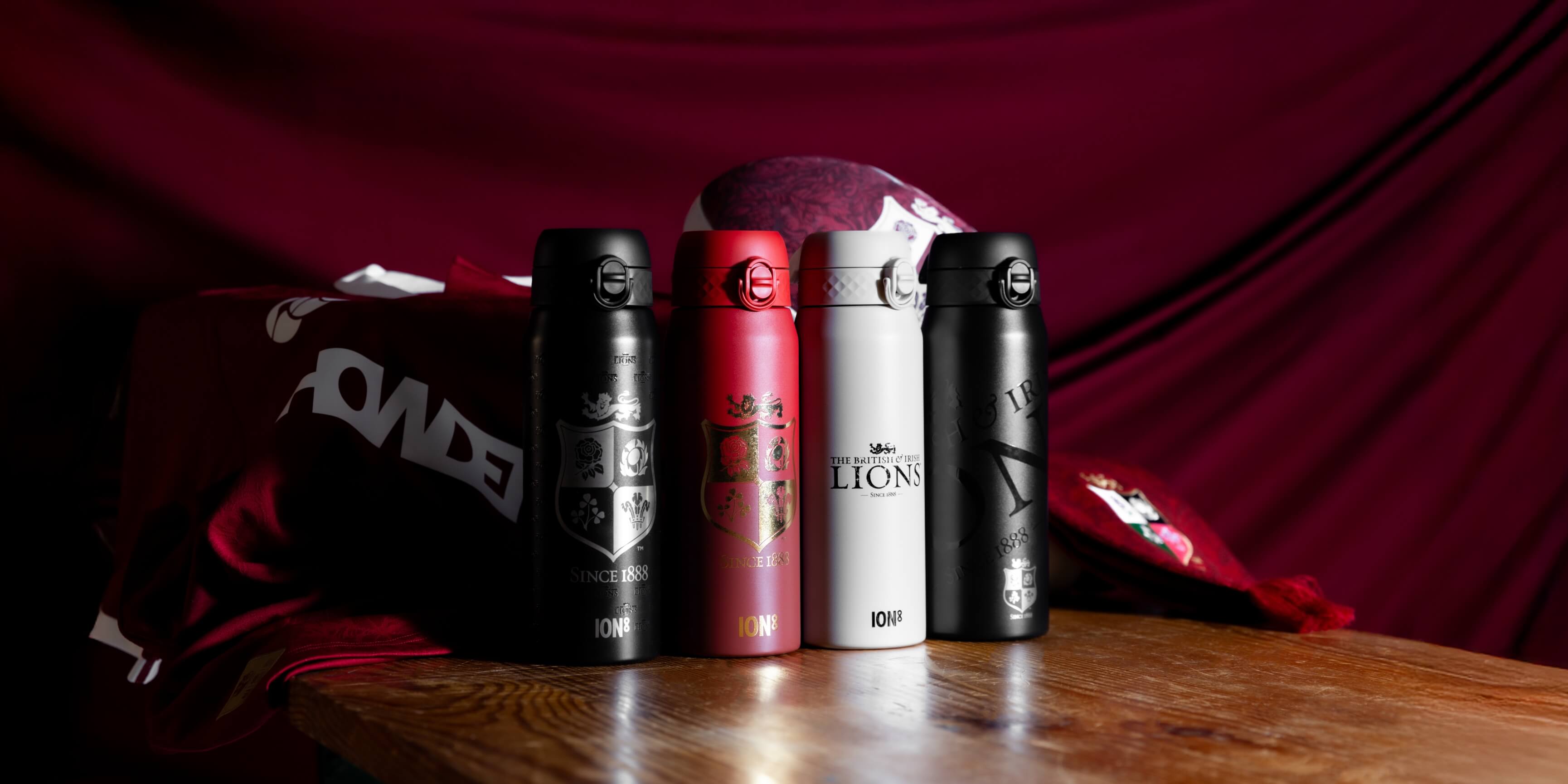 Four insulated water bottles, displaying British & Irish Lions logos, sit on a wooden table draped with a maroon cloth.