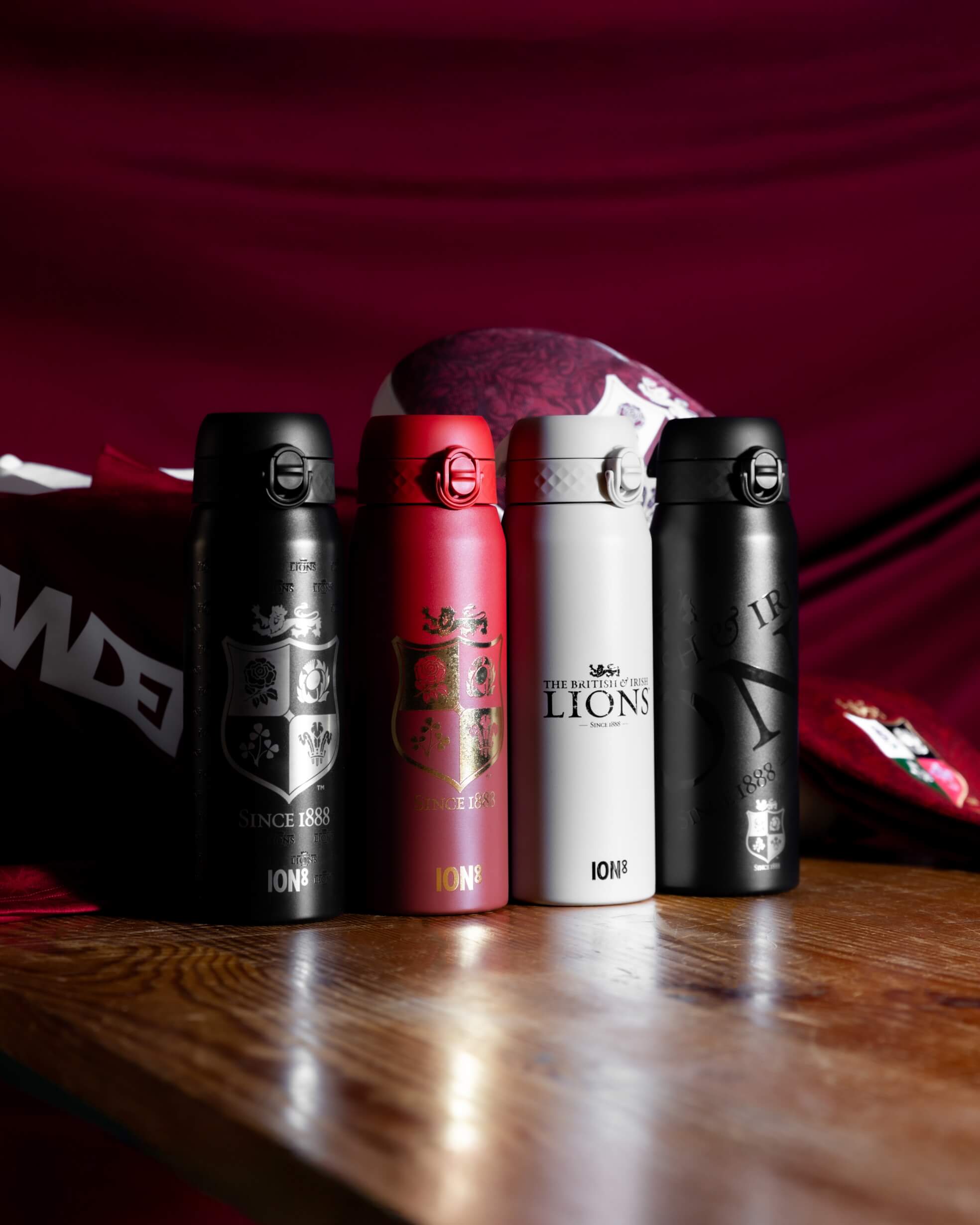 Four insulated bottles, displaying British and Irish Lions logos and "ION8" branding, sit on a wooden surface near a rugby jersey and ball.